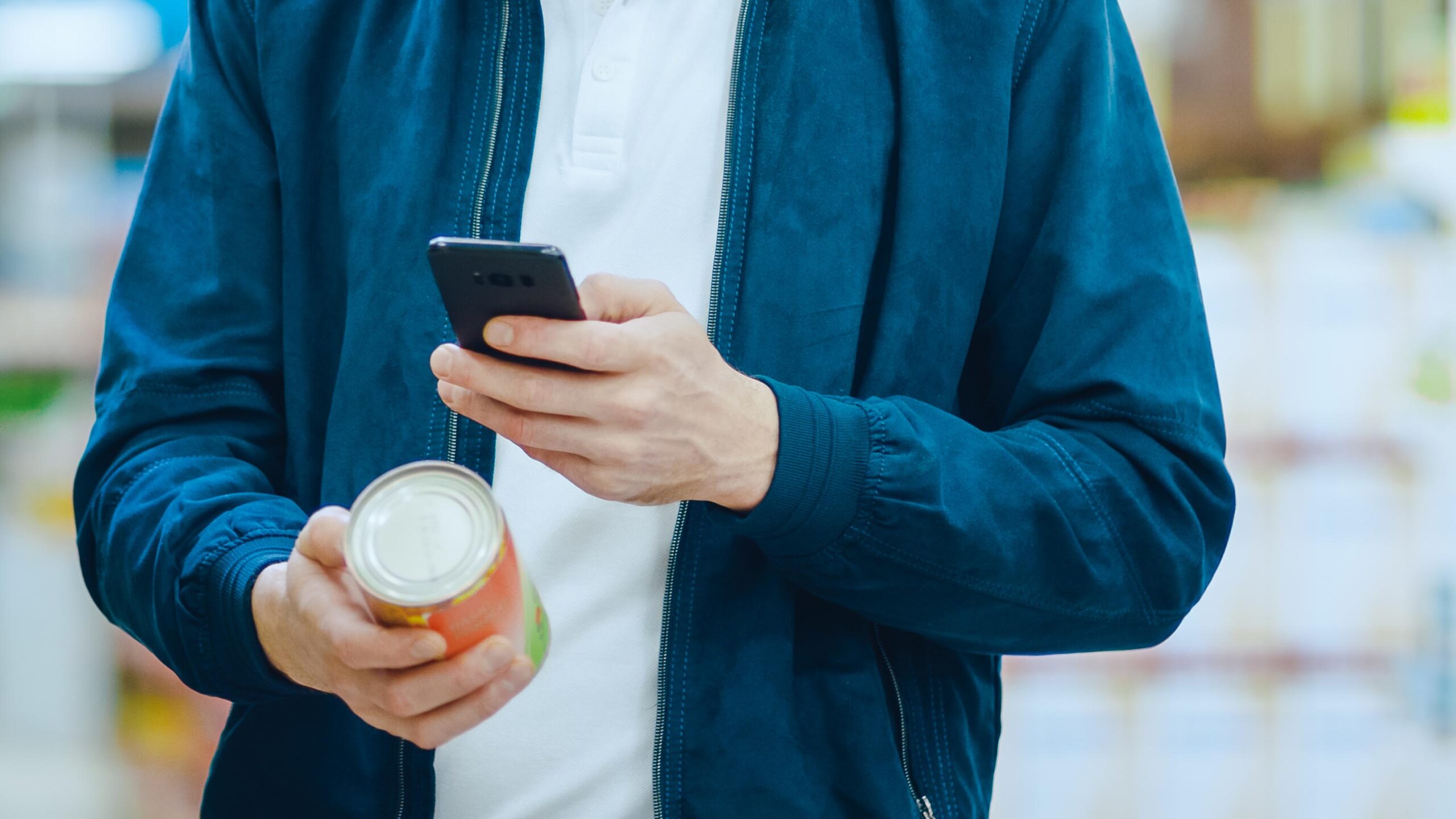 At the Supermarket: Handsome Man Uses Smartphone to Check Nutritional Value of the Canned Goods and Buy it. He's Standing with Shopping Cart in Canned Goods Section. Label cans